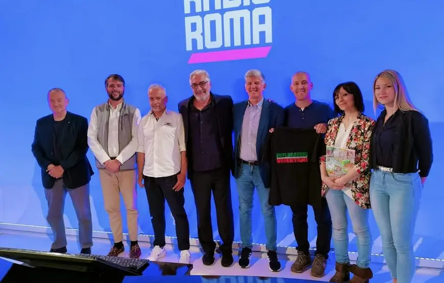 otto-persone-uomini-e-donne-sono-in-piedi-uno-di-fianco-all-atro-in-posa-per-la-fotografia-alle-loro-spalle-una-parete-blu-con-la-scritta-radio-roma-in-bianco